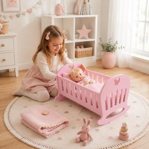 May include: A light pink wooden doll cradle with heart-shaped cutouts on the headboard and footboard. The cradle contains a doll with blonde hair wearing a white outfit, resting on a pink patterned blanket. A folded pink blanket sits nearby.