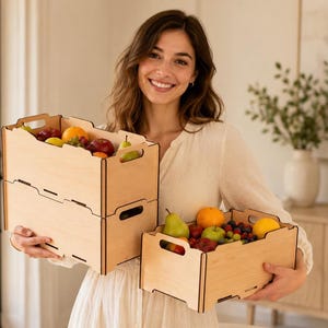 Puede incluir: Tres cajas rectangulares de madera de color claro con asas recortadas en los laterales. Dos cajas están apiladas verticalmente a la izquierda y una caja se sostiene a la derecha. Las cajas están llenas de varias frutas frescas, incluyendo manzanas, peras y naranjas.