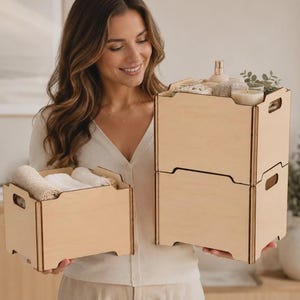 May include: Three light-coloured wooden storage crates with cutout handles. One smaller crate holds folded white textiles, while two larger crates are stacked vertically, containing a glass candle, a spray bottle, and greenery.