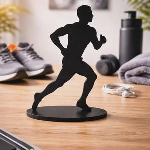 May include: A black metal silhouette of a runner in a sprinting pose, mounted on a circular base. The figure depicts a person in motion with arms bent and legs extended, capturing the dynamic posture of a runner in a solid, matte black finish.