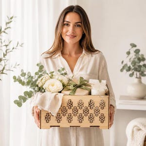 Puede incluir: Una caja de madera rectangular con un patrón geométrico repetitivo cortado con láser en su panel frontal. La caja contiene un ramo de flores blancas con hojas de eucalipto verdes y un pequeño regalo envuelto en papel marrón con un lazo.