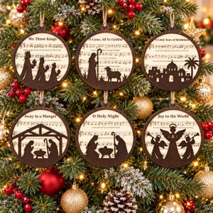 Könnte beinhalten: Sechs kreisförmige Weihnachtsanhänger aus Holz an einem Baum. Jeder Anhänger zeigt eine dunkelbraune, lasergeschnittene Silhouette einer Krippenszene oder religiösen Figur auf einem hellen Hintergrund mit aufgedruckten Notenblättern.