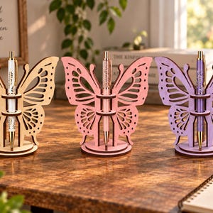May include: Three laser-cut wooden butterfly pen holders in light beige, soft pink, and light purple. Each butterfly features intricate wing cutouts and a central vertical stand with two circular rings to hold a matching colored pen upright.