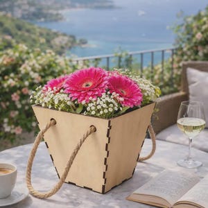 Peut inclure: Une jardinière carrée en bois avec des joints à entures multiples et des poignées en corde de jute épaisse, remplie de gerberas rose vif et de petites fleurs blanches de gypsophile. Le contenant a une finition en bois clair et une forme évasée.