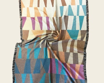 May include: Woven throw blanket with a geometric pattern in shades of brown, teal, purple, and peach. The blanket has a fringed edge in black and is folded to display the design.