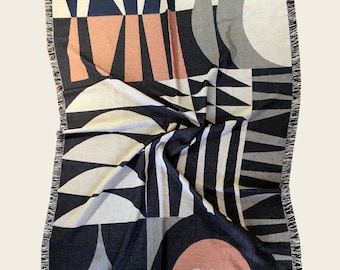 May include: A woven throw blanket with a geometric pattern in black, white, gray, and pink. The blanket has a fringed edge.