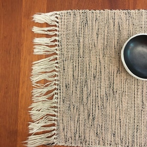 May include: A beige placemat with black stripes and white fringe. A black bowl is on the placemat.