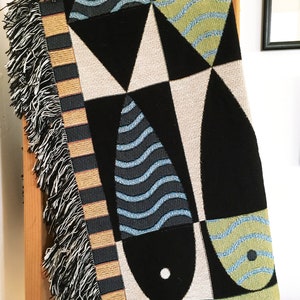 May include: A woven throw blanket with a geometric pattern of black, white, green, and blue. The pattern features stylized fish shapes.