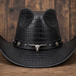 Op de afbeelding: Een zwarte cowboyhoed met een textuur van krokodillenleer. De hoed is voorzien van een zwarte band versierd met zilverkleurige metalen studs en een opvallend zilverkleurig embleem van een stierenschedel aan de voorzijde.