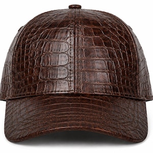 Puede incluir: Una gorra de béisbol de color marrón oscuro con un patrón de textura de piel de cocodrilo en toda la superficie. La gorra presenta una corona estructurada, una visera curva y un pequeño botón a juego en el centro superior de la cúpula.