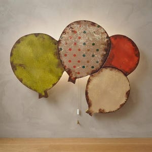 Pode incluir: Uma luminária de parede em forma de um cacho de quatro balões. Os balões apresentam texturas e cores distintas, incluindo um verde-limão texturizado, um bege com bolinhas vermelhas e verdes, um vermelho-alaranjado sólido e um balão bege liso.