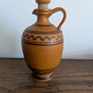 Puede incluir: Una vinagrera de cerámica color amarillo mostaza con acabado de esmalte craquelado, que presenta bandas horizontales y un patrón de líneas onduladas en marrón oscuro. El recipiente tiene un asa, un pico estrecho y un tapón de cerámica con corcho.