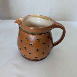 Pode incluir: Uma pequena leiteira de cerâmica castanha, de forma arredondada com acabamento brilhante. O exterior apresenta um padrão repetitivo de pequenos pontos escuros dispostos em grupos. Possui uma asa curva, uma borda larga e um bico vertedor.