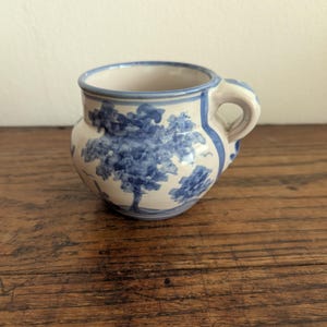 Könnte beinhalten: Ein kleiner Keramikbecher mit bauchigem Körper und einem einzelnen Henkel. Der Becher hat eine cremefarbene Basis mit handgemalten blauen Motiven, die Bäume und Blattwerk darstellen. Ein blauer Streifen verläuft um den Rand und den Henkelansatz.
