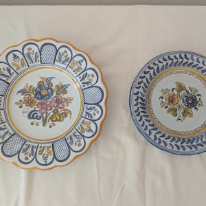 May include: Two ceramic plates featuring hand-painted floral motifs in blue, yellow, and green. The left plate has a scalloped edge with a lattice pattern, while the right plate features a repeating leaf border design around a central flower bouquet.
