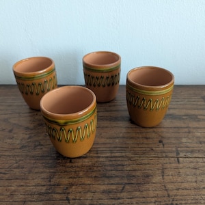 Può includere: Tre tazze in ceramica color terracotta con finitura liscia. Ogni tazza presenta una fascia orizzontale verde vicino al bordo e un motivo a zigzag verticale ripetuto in verde attorno al corpo esterno. L'interno delle tazze non è smaltato.