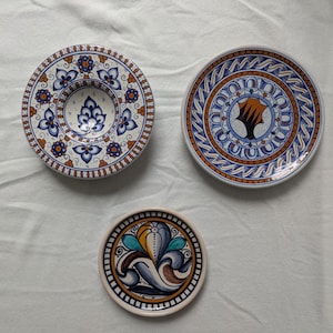 Può includere: Tre piatti in ceramica con disegni dipinti a mano. Il piatto in alto a sinistra ha un centro profondo con motivi floreali blu e arancioni. Il piatto in alto a destra mostra un motivo centrale a stella arancione. Motivo floreale stilizzato.