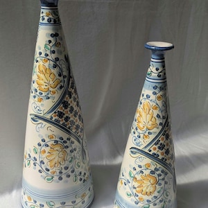 Puede incluir: Dos jarrones cónicos de cerámica con base color crema, decorados con motivos florales pintados a mano en azul y amarillo, enredaderas intrincadas y patrones de cuadrícula geométricos. Cada jarrón tiene un cuello estrecho y un borde circular.