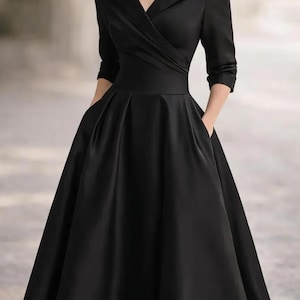 May include: A black midi dress featuring a wide shawl collar, a wrap-style bodice with subtle pleating, three-quarter length sleeves, and a full, pleated skirt with side pockets. The dress is paired with a silver-toned crystal necklace.