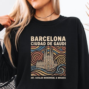 May include: A black long-sleeve sweatshirt featuring a graphic print of the Sagrada Familia in Barcelona. The design includes swirling mosaic patterns in blue, orange, and tan, with the text "BARCELONA CIUDAD DE GAUDI" and "ART, CATALAN MODERNISM, & MOSAICS."