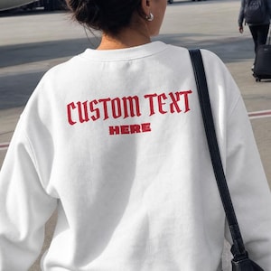 May include: A white long-sleeve crewneck sweatshirt featuring the red text CUSTOM TEXT HERE printed on the upper back. The garment has a ribbed neckline and cuffs, and is worn with a black shoulder bag and dark grey trousers.