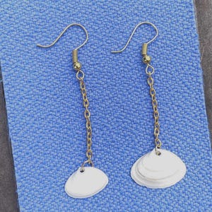 Può includere: Un paio di orecchini pendenti con ganci in metallo dorato e sottili catenelle a maglie. Ogni orecchino è rifinito con un piccolo ciondolo a forma di conchiglia bianca e liscia alla base, creando un design semplice ed equilibrato per questi gioielli.