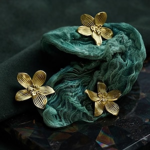 May include: Three gold-toned metal flower-shaped ornaments with five petals each, featuring a ribbed texture and a central stamen detail. The flowers are arranged on a piece of dark teal, textured, semi-sheer fabric with a gathered, layered appearance.