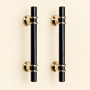 May include: Two cylindrical door handles featuring a glossy black finish. Each handle is secured by two polished gold-tone metal mounts at the top and bottom, which connect the black bar to circular wall plates with a reflective metallic surface.