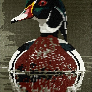 May include: A cross-stitch embroidery pattern featuring a wood duck with a dark green, black, and white head, a red eye, and a white beak. The chest is a deep reddish-brown color, set against a textured, dark olive green background grid.