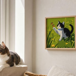 May include: A square wall art piece in a light wood frame featuring a pixelated illustration of a black and white cat with a blue collar reaching toward a small orange butterfly in a field of green grass and yellow flowers.