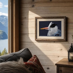 May include: A framed needlepoint artwork depicting a grey heron in profile against a dark background. The bird features a white crest, a long orange beak, and detailed grey plumage. The artwork is enclosed in a textured brown wooden frame.