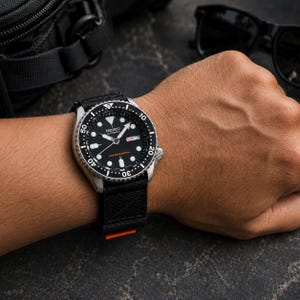 Puede incluir: Un reloj de buceo automático Seiko con esfera negra, marcadores de hora luminosos y bisel giratorio. El reloj cuenta con una ventana de día y fecha en la posición de las tres, manecillas plateadas y una correa de nailon negra con detalle naranja.