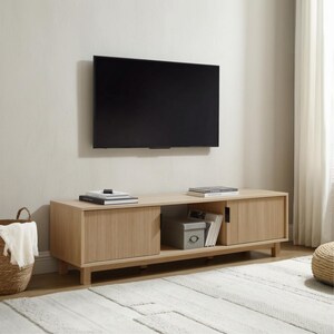 May include: A light wood media console featuring two sliding doors with a vertical ribbed texture. The unit has an open central shelf containing a grey storage box and books, with a flat-screen television mounted on the wall directly above it.