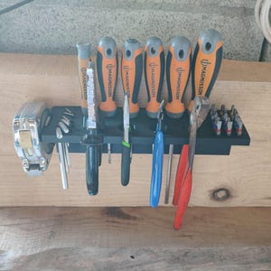 Op de afbeelding: Een zwarte wandhouder voor gereedschap met diverse gereedschappen, waaronder een zilveren rolmaat, verschillende Magnusson-schroevendraaiers met oranje en grijze handvatten, een punttang, een blauwe tang, een rode tang en metalen bitjes.
