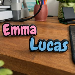 Könnte beinhalten: Zwei 3D-gedruckte Namensschilder mit schwarzem Rand. Ein Schild zeigt den Namen Emma in rosa Buchstaben, das andere Schild zeigt den Namen Lucas in hellblauen Buchstaben. Beide Schilder haben eine abgerundete, fette Schriftart auf einer Holzoberfläche.