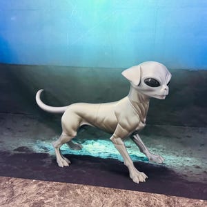 May include: A light gray, articulated figurine of a dog with an alien-like head, featuring large, smooth, black, almond-shaped eyes, pointed ears, and a slender, muscular body with a curved tail, standing in a profile position on a flat surface.