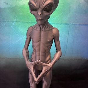 May include: A grey, humanoid alien figurine with a large, smooth, elongated head, dark almond-shaped eyes, and a slender, muscular build. The figure stands in an upright position with its hands joined together in front of its torso.