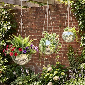 May include: Three spherical hanging planters covered in reflective silver mirror tiles, each suspended by silver metal chains. The planters contain various green foliage, including ferns, trailing vines, and small succulents, with red and purple flowers.