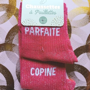 Peut inclure: Une paire de chaussettes rose vif à paillettes avec du texte blanc imprimé sur le tissu. La chaussette du haut porte le mot PARFAITE et celle du bas le mot COPINE. Les chaussettes sont fixées sur un carton avec le texte Chaussettes à Paillettes et une taille 36-42.