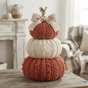 May include: A stack of three decorative pumpkins featuring textured knit patterns in burnt orange and cream colors. The top pumpkin is adorned with a beige fabric bow and two small birch wood sticks as a stem, creating a vertical arrangement.