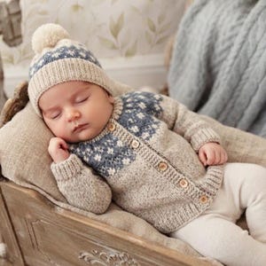 May include: A beige and blue knit cardigan with a fair isle pattern on the yoke and sleeves, featuring five wooden buttons down the front. The garment is paired with a matching beige and blue knit beanie hat with a pom-pom on the top.