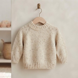 May include: A long-sleeved jumper in a light cream colour with a speckled pattern throughout. The garment features a ribbed crew neckline, ribbed cuffs, and a ribbed hem. It is displayed on a wooden hanger against a neutral-coloured wall.