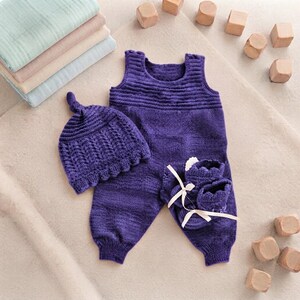 May include: A deep purple knitted set consisting of a sleeveless romper with a ribbed chest, a matching beanie with a top knot, and a pair of booties featuring white ribbon ties. The set is displayed alongside a stack of folded muslin cloths.