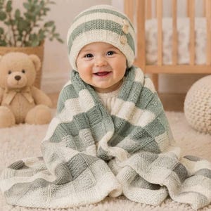Könnte beinhalten: Eine gestrickte Babymütze und eine passende Decke mit einem Karomuster in Salbeigrün und Creme. Die Mütze ist an der Seite mit drei kleinen Holzknöpfen versehen. Das Set ist in einem strukturierten Strickmuster gefertigt und wirkt weich und gleichmäßig.
