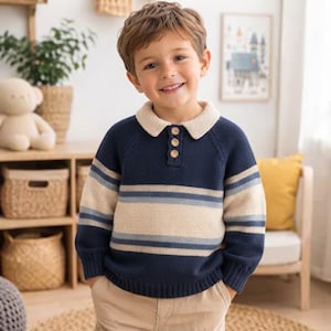 May include: A long-sleeve knit jumper featuring a navy blue base with a wide beige horizontal stripe across the chest and sleeves, accented by thin light blue stripes. The jumper has a beige collar and a three-button placket at the neck.