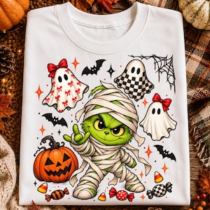 May include: A white t-shirt featuring a central green character wrapped in bandages like a mummy, surrounded by three ghosts, a jack-o'-lantern, bats, candy corn, and wrapped candies. The ghosts have red bows or checkered patterns.