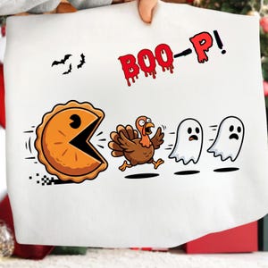 May include: A white t-shirt featuring a graphic of an orange pie shaped like a video game character chasing a brown turkey and two white ghosts. The text BOO-P! is printed in red with a dripping effect above the characters, alongside three black bats.