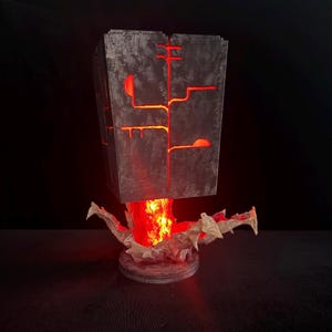 May include: A rectangular gray lamp with a textured surface featuring cutouts that emit a bright red light. The lamp is supported by a jagged, rock-like base that also glows with an intense red light, creating a visual effect of energy.