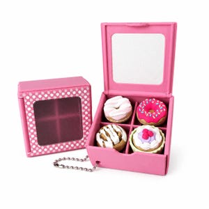 May include: A pink square box with a checkered lid and a mirror, containing four miniature food-shaped erasers. The erasers include a white-frosted donut, a pink-frosted donut with sprinkles, a cinnamon roll, and a cupcake with berries.