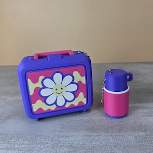 May include: A purple rectangular lunchbox with a pink handle and a central panel featuring a white daisy with a yellow smiley face center against a wavy yellow and pink background, accompanied by a small purple and pink cylindrical container.